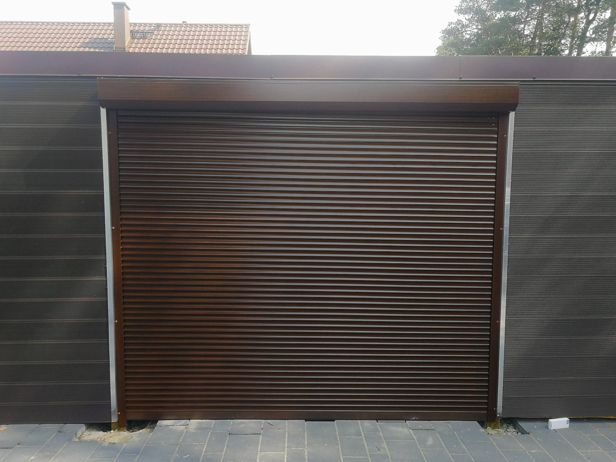 Zewnętrzna, brązowa roleta zabezpieczająca, z widoczną fakturą poziomych lameli, zamontowana na tle elewacji z szarego panelu kompozytowego.