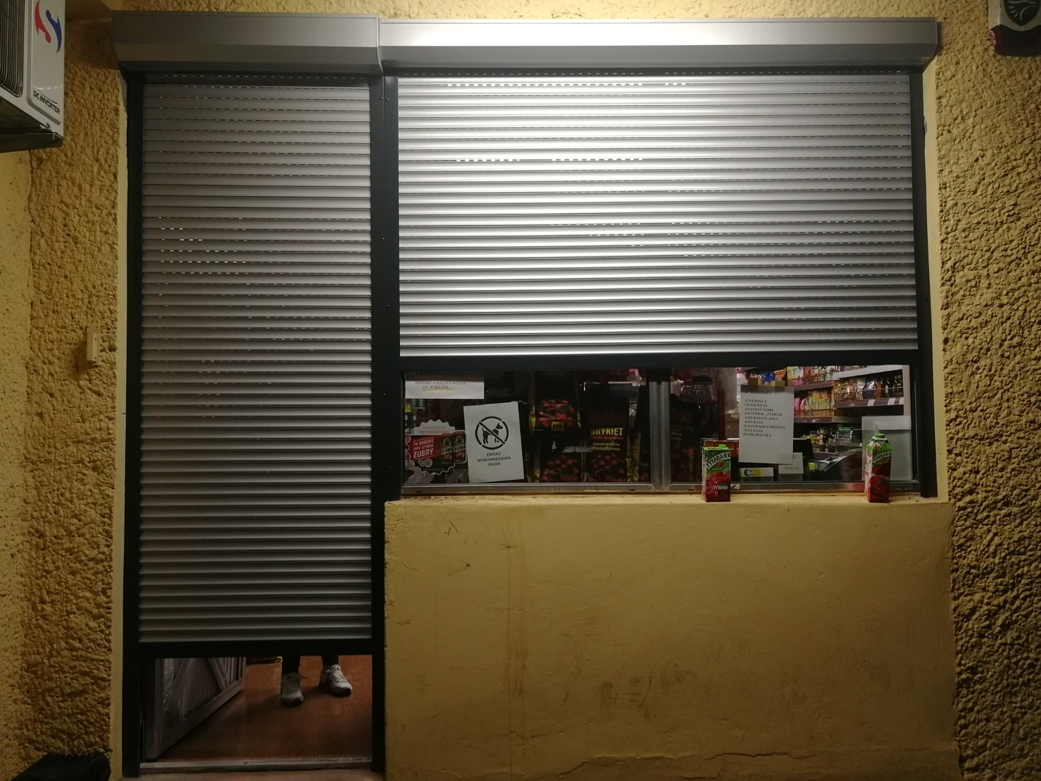Zewnętrzna, metalowa roleta antywłamaniowa częściowo opuszczona na okno małego sklepu, widoczne produkty wewnątrz i fragment nóg osoby wchodzącej do sklepu.