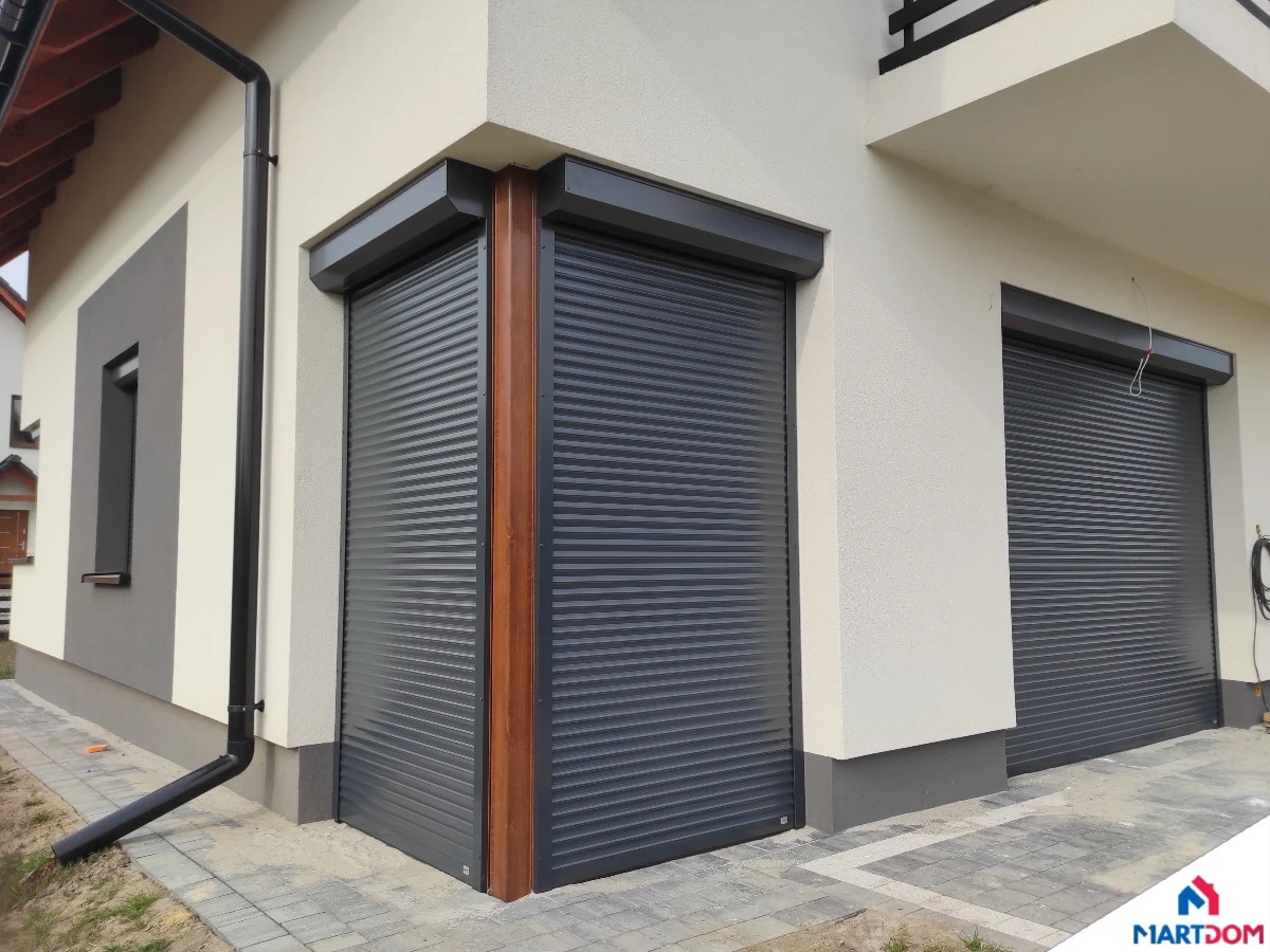 Elewacja domu z dwiema szarymi roletami zewnętrznymi, jedna z drewnianą ramą, widoczne rury spustowe i kostka brukowa.