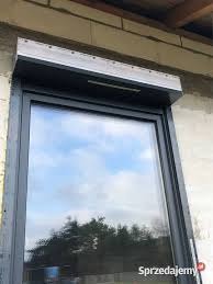 Zamknięta roleta zewnętrzna w szarej ramie okiennej, odbicie nieba i drzew w szybie, surowy mur budynku w tle.