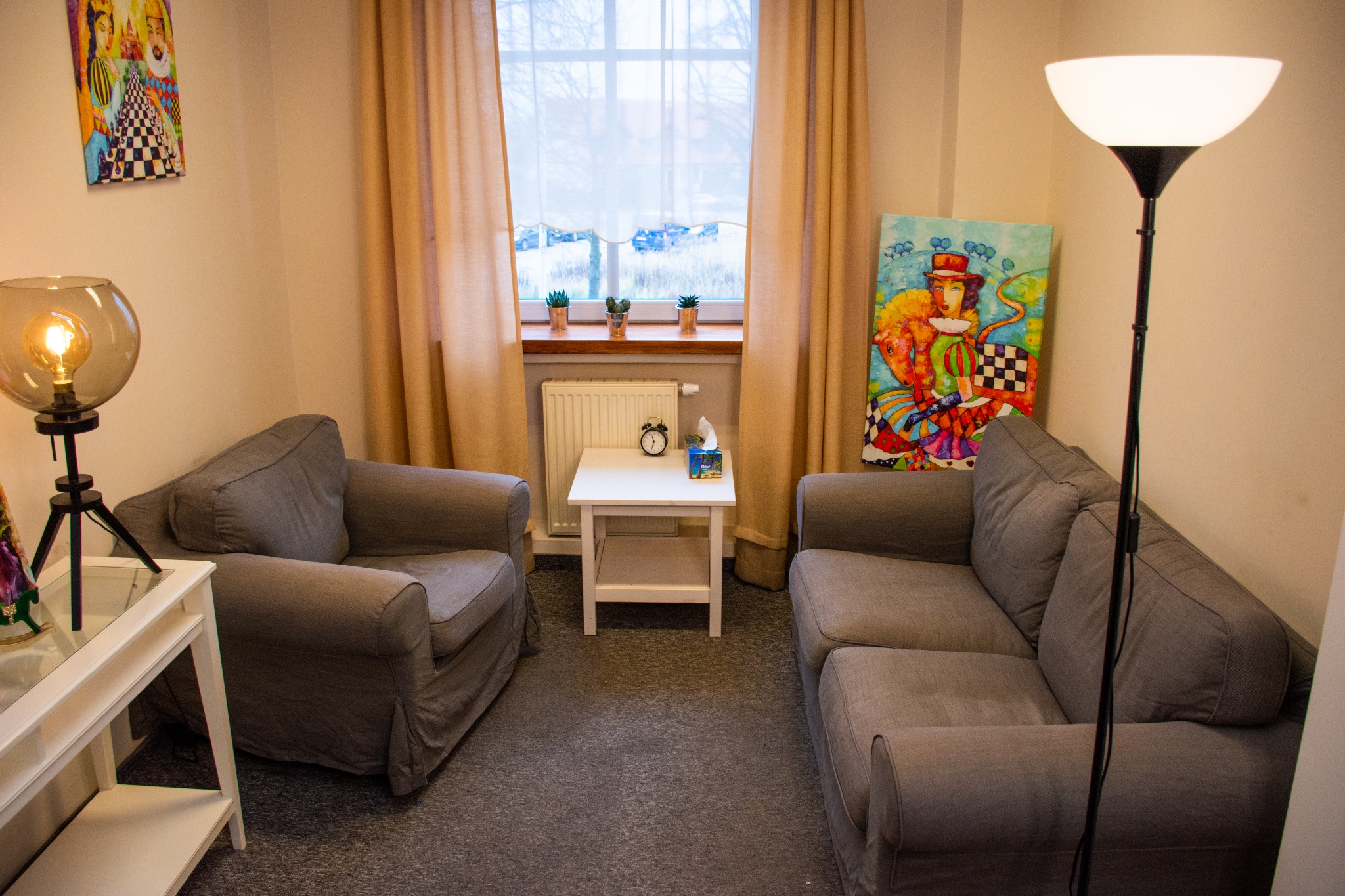 Przytulny gabinet z dwoma szarymi fotelami, sofą, białym stolikiem z zegarem i chusteczkami, lampą stojącą i obrazami na ścianach, oświetlony światłem dziennym wpadającym przez okno z trzema...