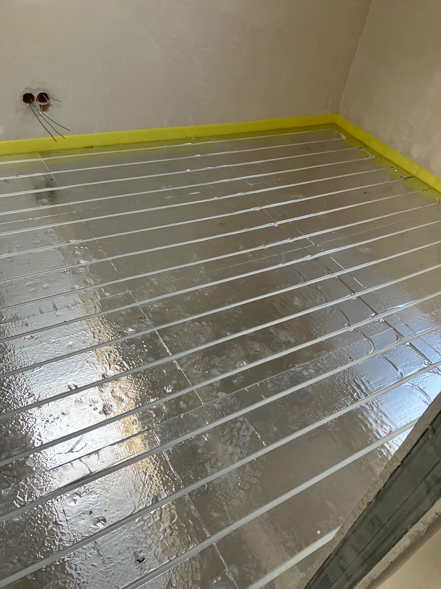 Rozłożona folia aluminiowa z rurkami ogrzewania podłogowego, żółta taśma brzegowa, surowe ściany i wystające kable elektryczne.