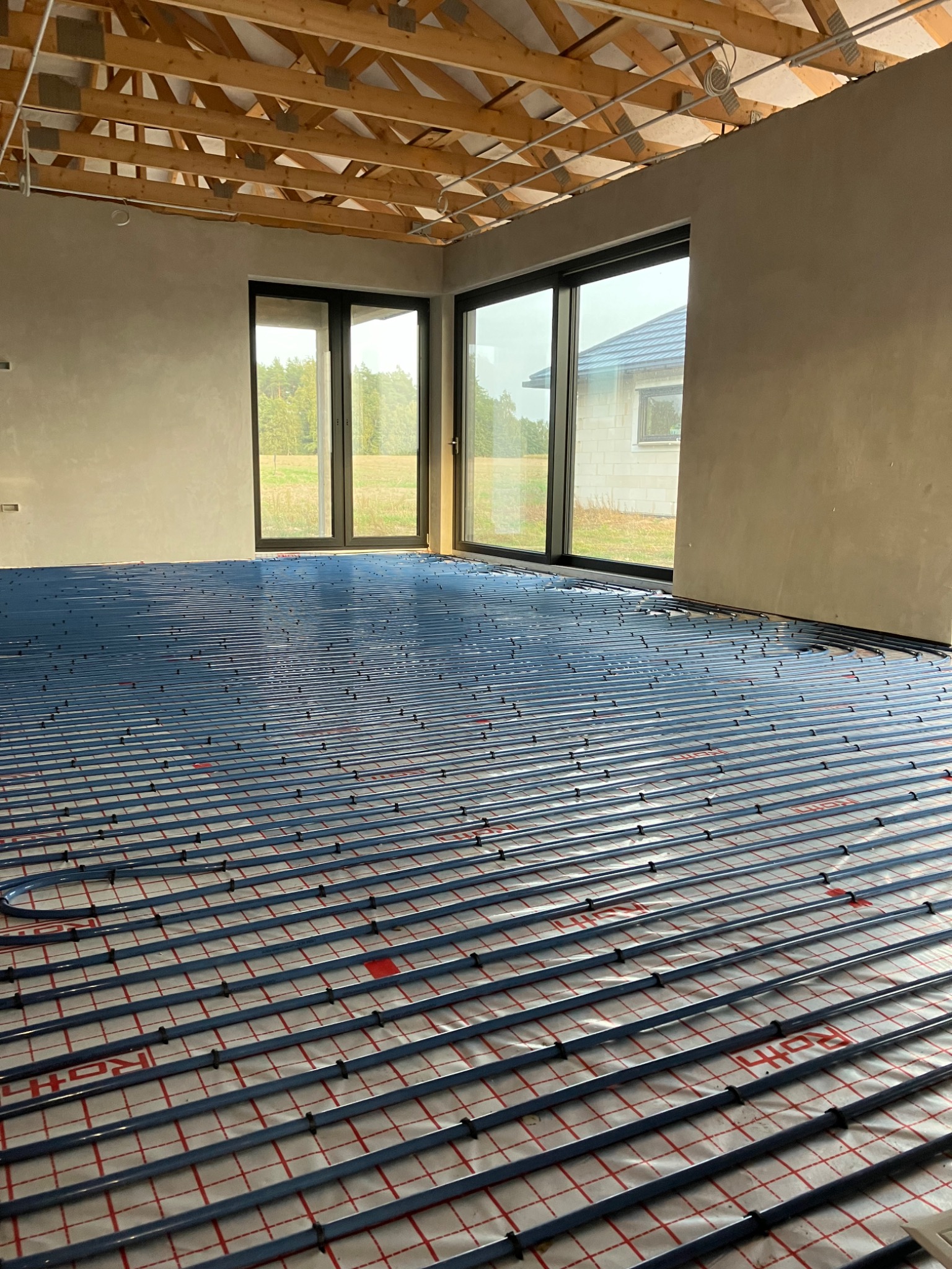 Niebieskie rury ogrzewania podłogowego Roth ułożone na izolacji z czerwoną siatką w surowym wnętrzu z dużymi oknami i widoczną konstrukcją dachu.