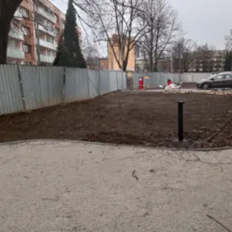 Teren przygotowany pod nasadzenia, ogrodzony blachą falistą, z widocznymi paletami i elementami małej architektury w tle zabudowa miejska.