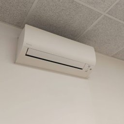 Clean&KlimaKing - Biała jednostka klimatyzacji Daikin zamontowana na ścianie pod sufitem kasetonowym w jasnym pomieszczeniu biurowym.