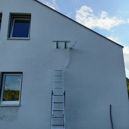 Clean&KlimaKing - Montaż wspornika klimatyzatora na elewacji domu jednorodzinnego, widoczna drabina oparta o ścianę, przewody i uchwyt na tle jasnego nieba z chmurami.