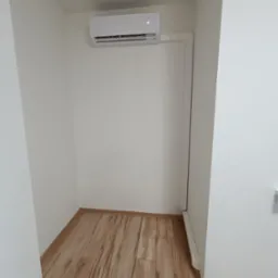 Biały klimatyzator ścienny zamontowany w niewielkim, pomalowanym na biało pomieszczeniu z panelami podłogowymi imitującymi drewno.