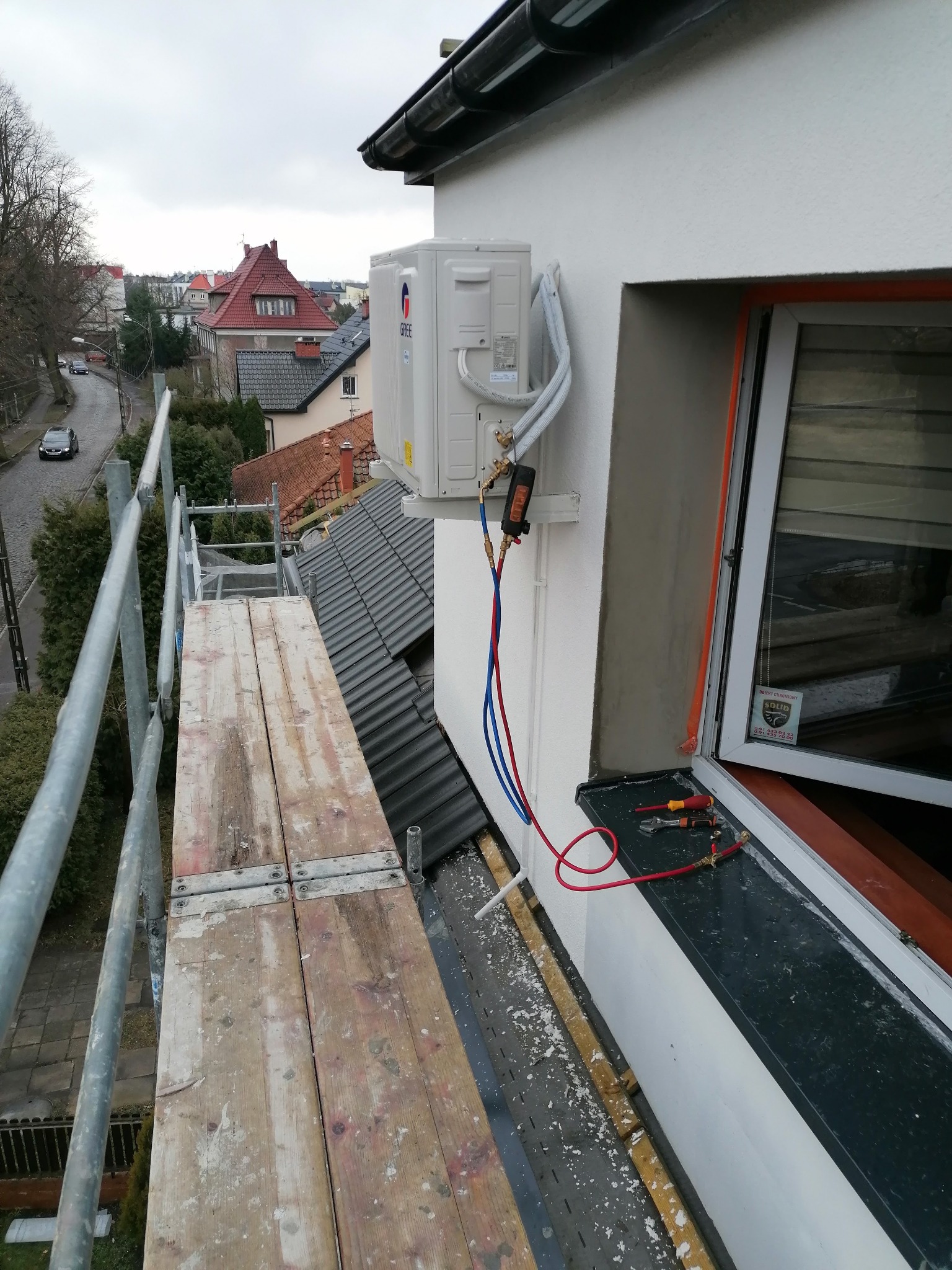 Montaż zewnętrznej jednostki klimatyzatora marki Gree na elewacji budynku, widok z rusztowania, podłączone przewody serwisowe, narzędzia leżą na parapecie okiennym.