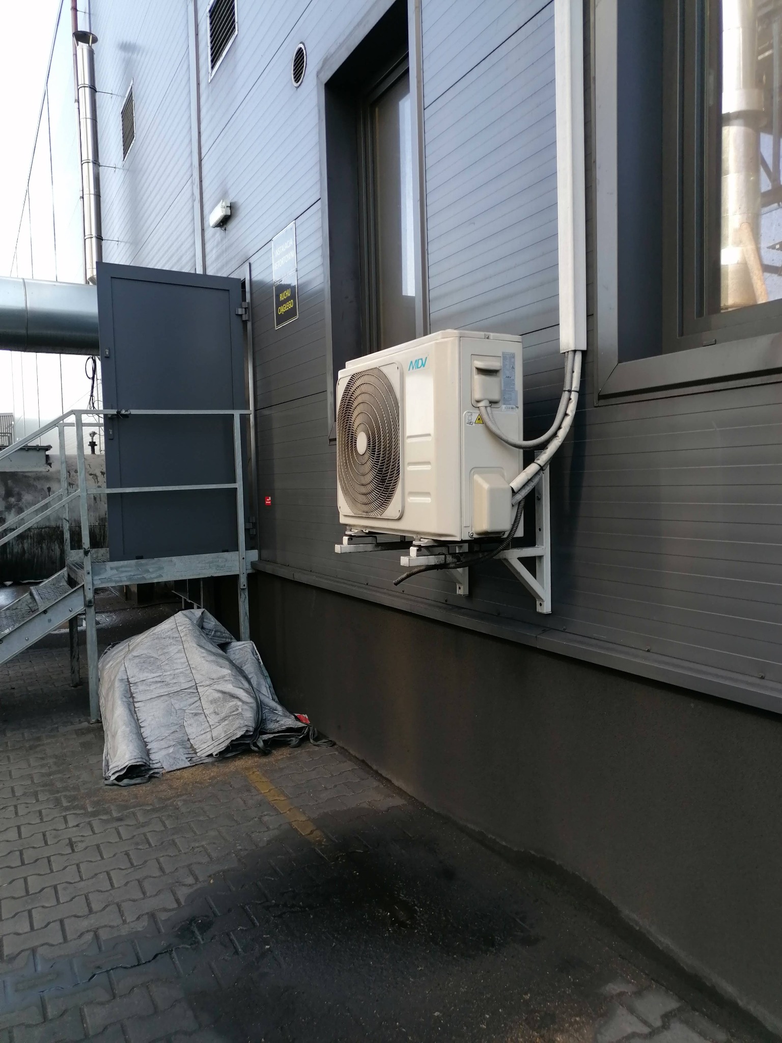 Zewnętrzna jednostka klimatyzacji MDV zamontowana na szarej, metalowej ścianie budynku przemysłowego, widoczne przewody i wsporniki montażowe, obok wejście z metalowymi schodami, pod spodem plandeka.