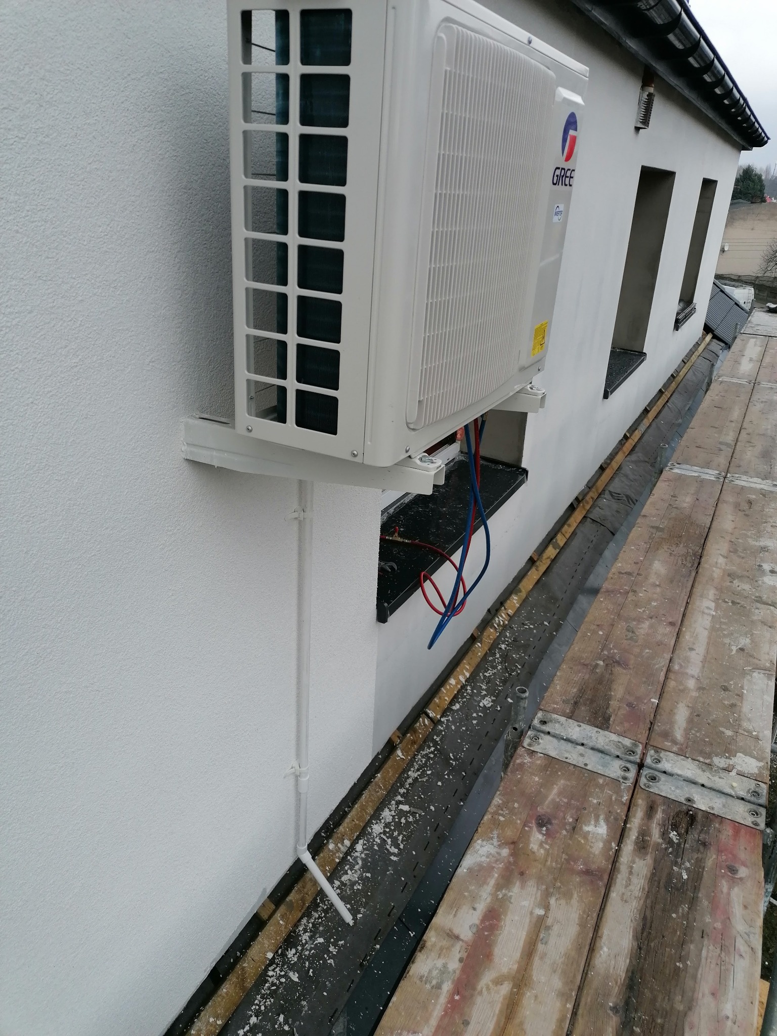 Biały klimatyzator marki Gree zamontowany na ścianie budynku z widocznymi przewodami, ujęcie z rusztowania.