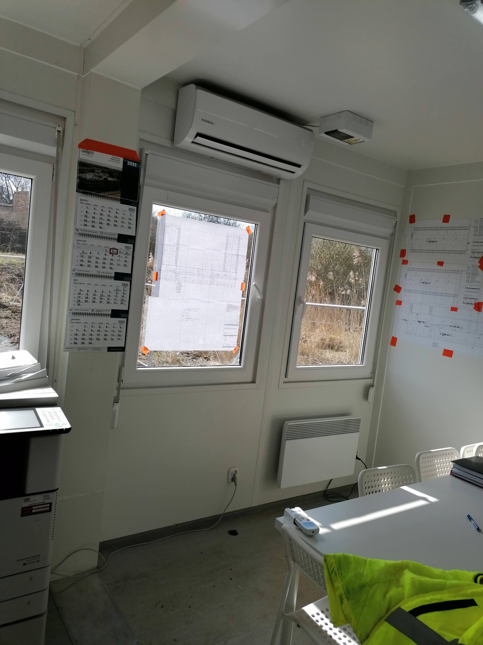 Wnętrze biura kontenerowego z zamontowaną klimatyzacją Kaisai, kalendarzem na ścianie i planami budowlanymi przyklejonymi taśmą do okna.