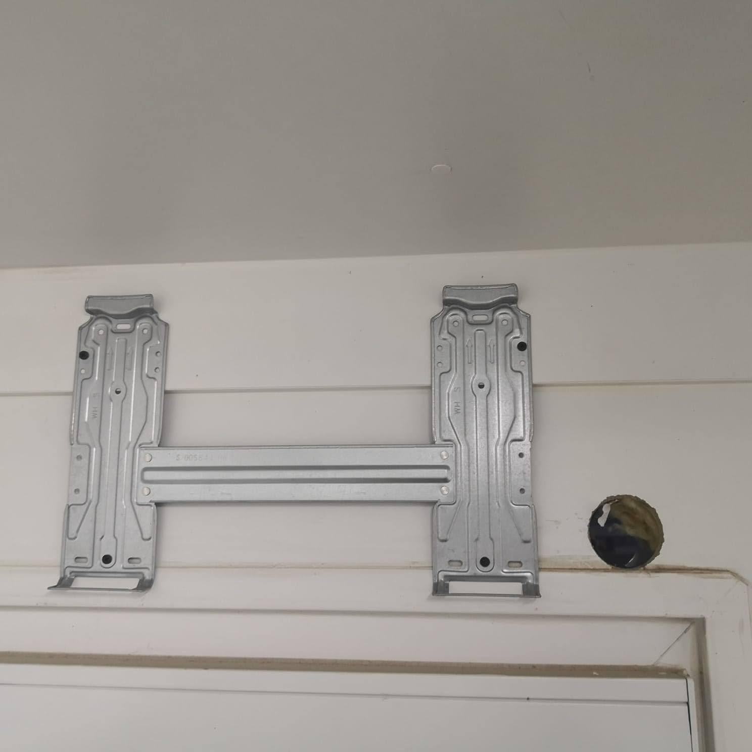 Metalowy wspornik montażowy klimatyzatora przymocowany do białej ściany z widocznym otworem na przewody.