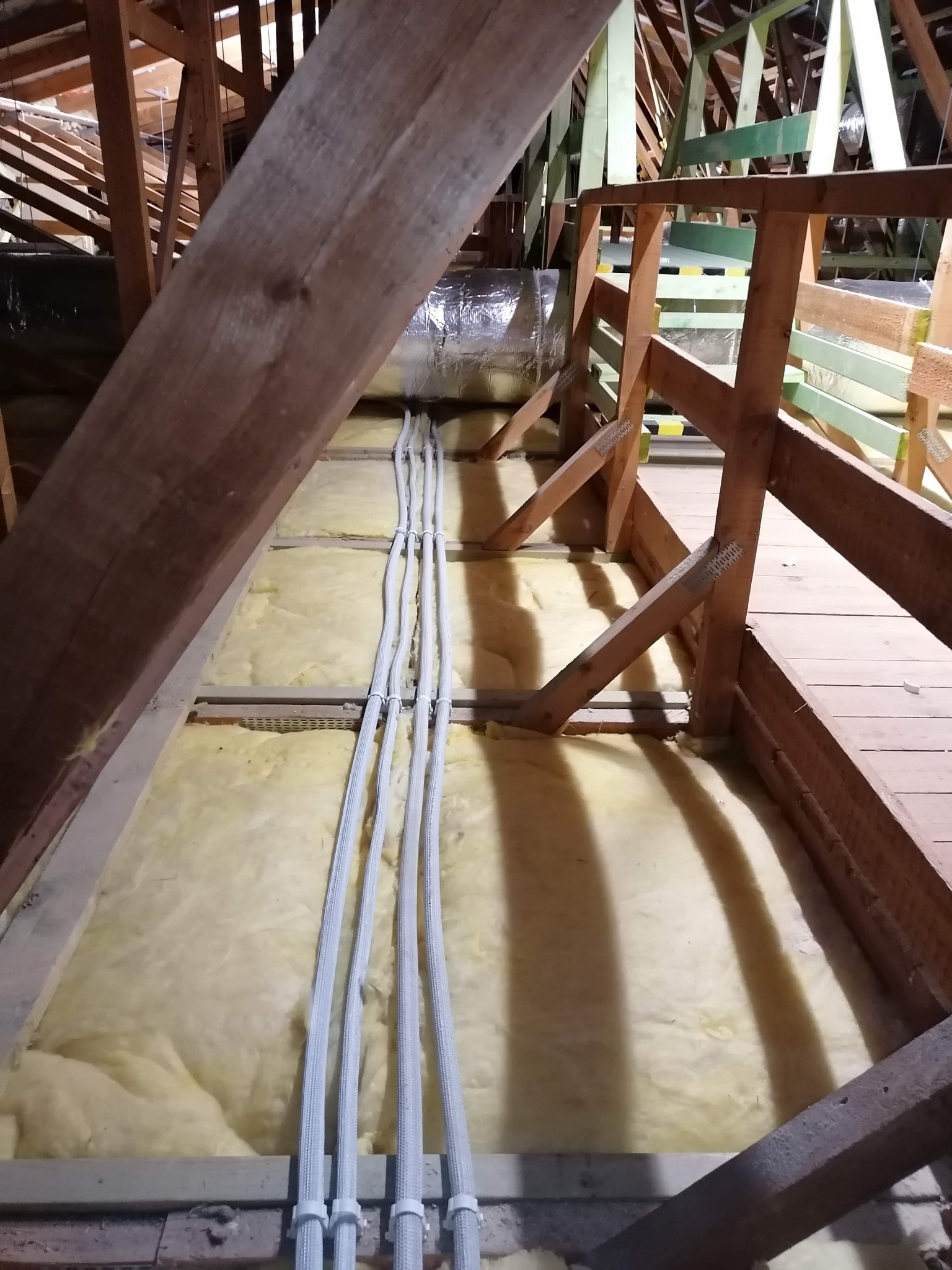 Instalacja przewodów w izolowanej wełną mineralną przestrzeni poddasza, widoczne drewniane elementy konstrukcyjne dachu i rura wentylacyjna w tle.