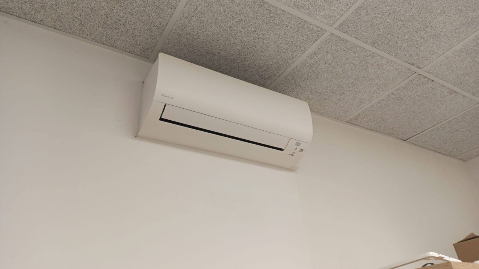 Biała jednostka klimatyzacji Daikin zamontowana na ścianie pod sufitem kasetonowym w jasnym pomieszczeniu biurowym.