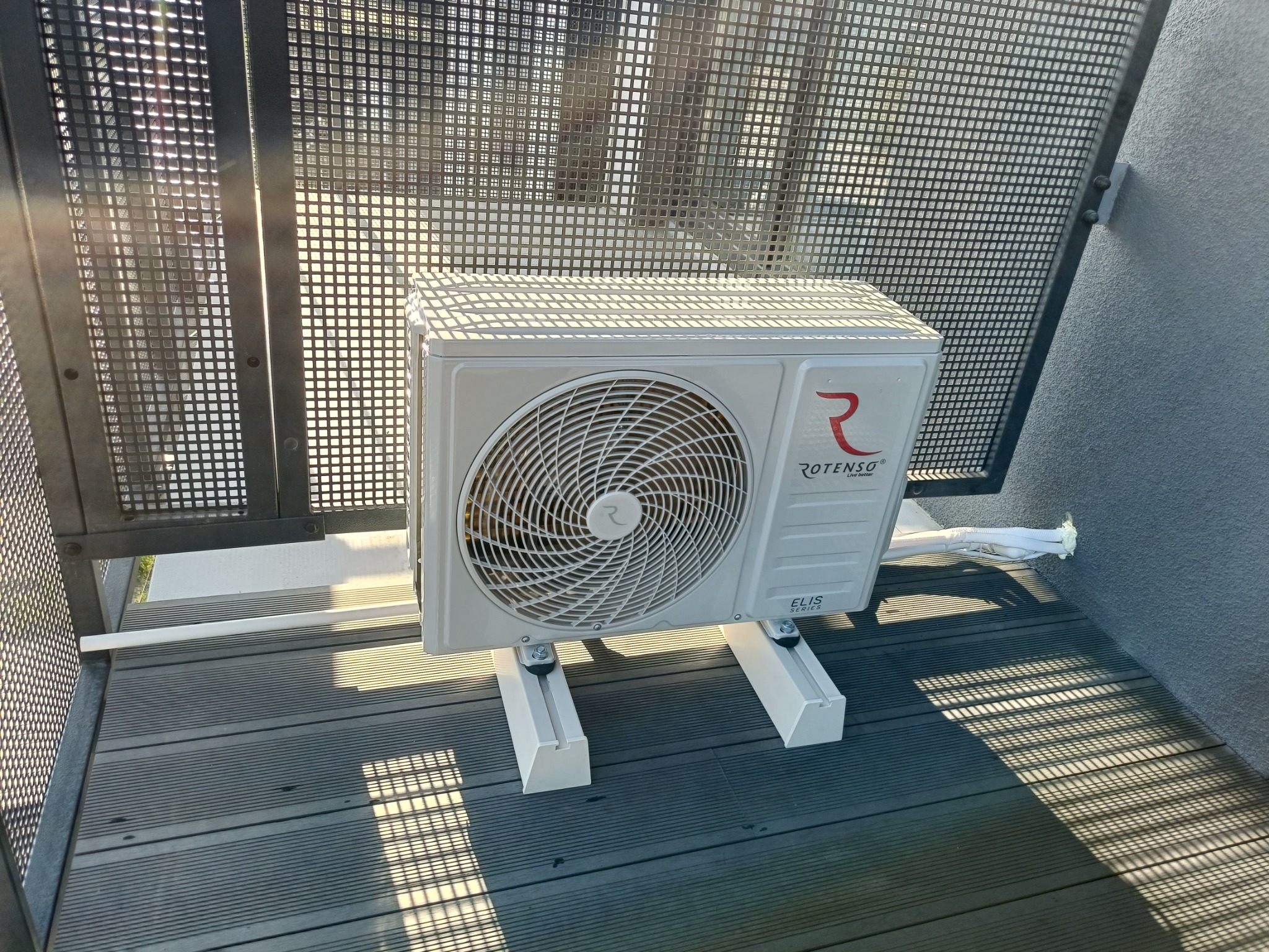 Zewnętrzna jednostka klimatyzatora Rotenso ELIS Series, zamontowana na wspornikach na tarasie z desek kompozytowych, z widocznymi przewodami odprowadzającymi skropliny i metalową kratą w tle.