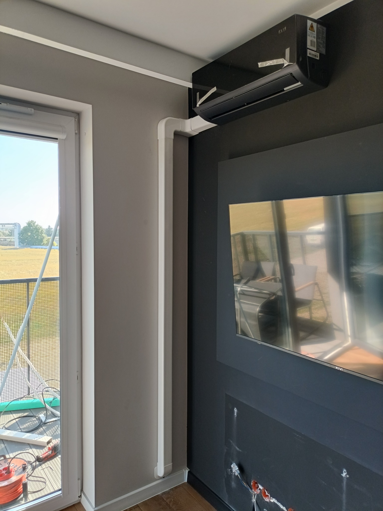 Czarny klimatyzator ścienny zamontowany w pokoju, z widocznymi białymi listwami maskującymi instalację, częściowo zakryty przez telewizor, z widokiem na balkon i pole za oknem.