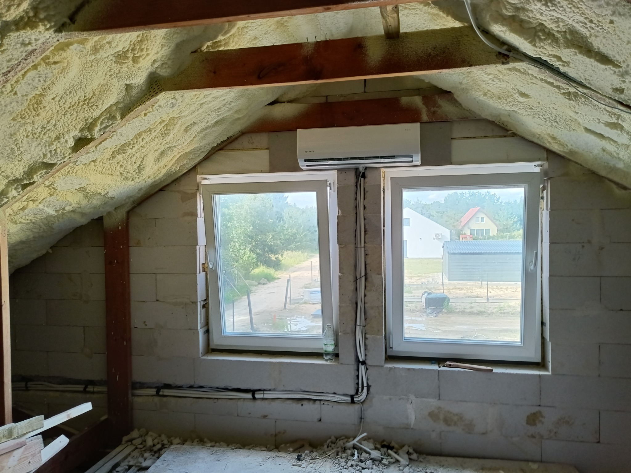 Montaż klimatyzatora na poddaszu domu w trakcie wykańczania, widoczne okna, ocieplenie pianką, instalacja elektryczna.