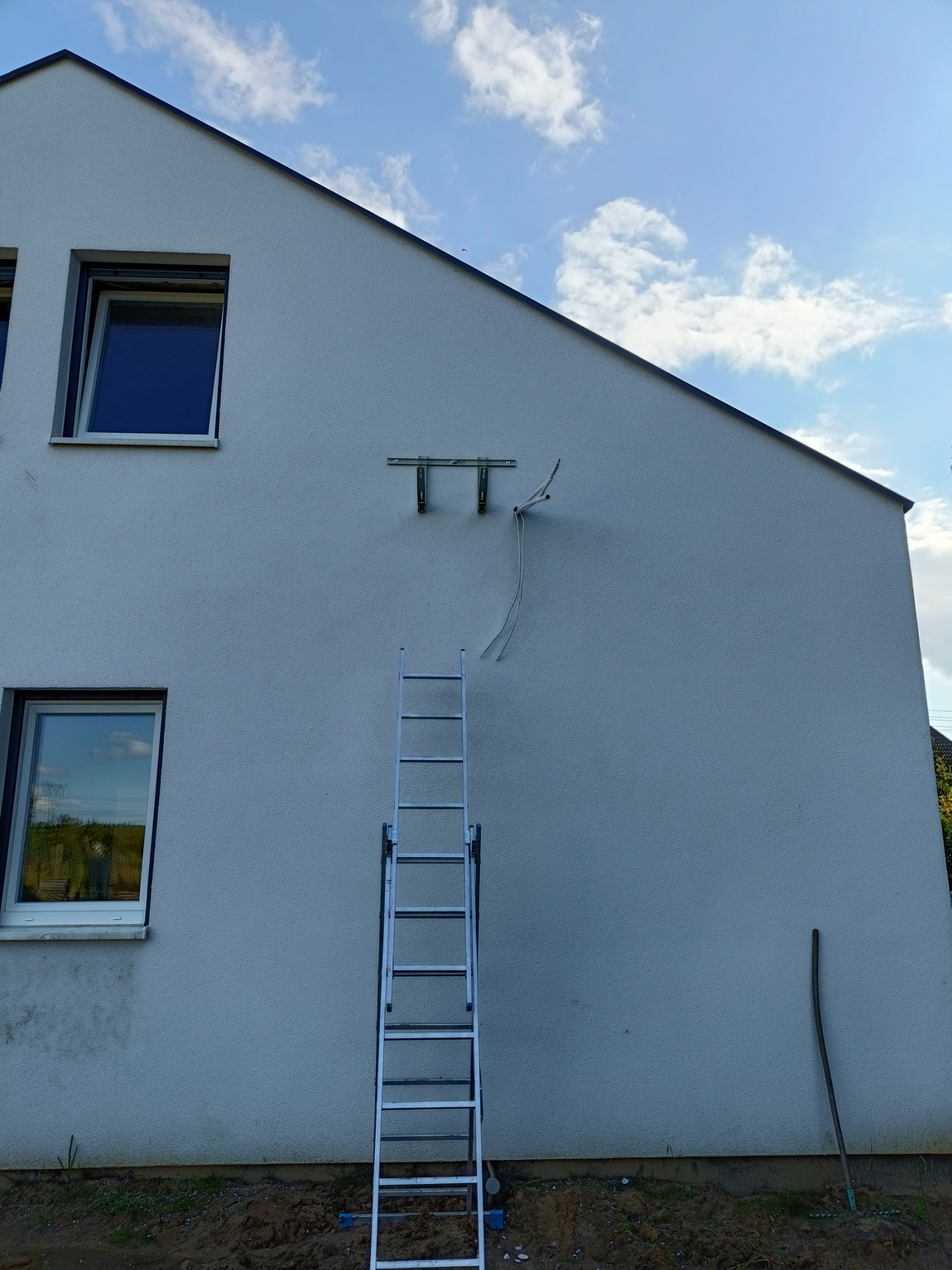 Montaż wspornika klimatyzatora na elewacji domu jednorodzinnego, widoczna drabina oparta o ścianę, przewody i uchwyt na tle jasnego nieba z chmurami.
