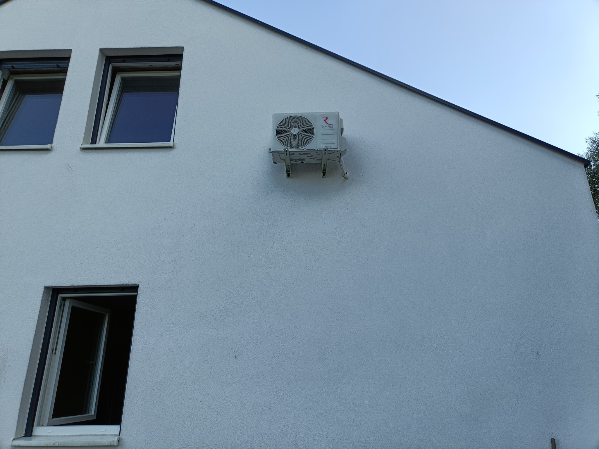Biała ściana budynku z zamontowaną jednostką zewnętrzną klimatyzacji Rotenso, widoczne dwa okna, jedno otwarte, niebieskie niebo w tle.