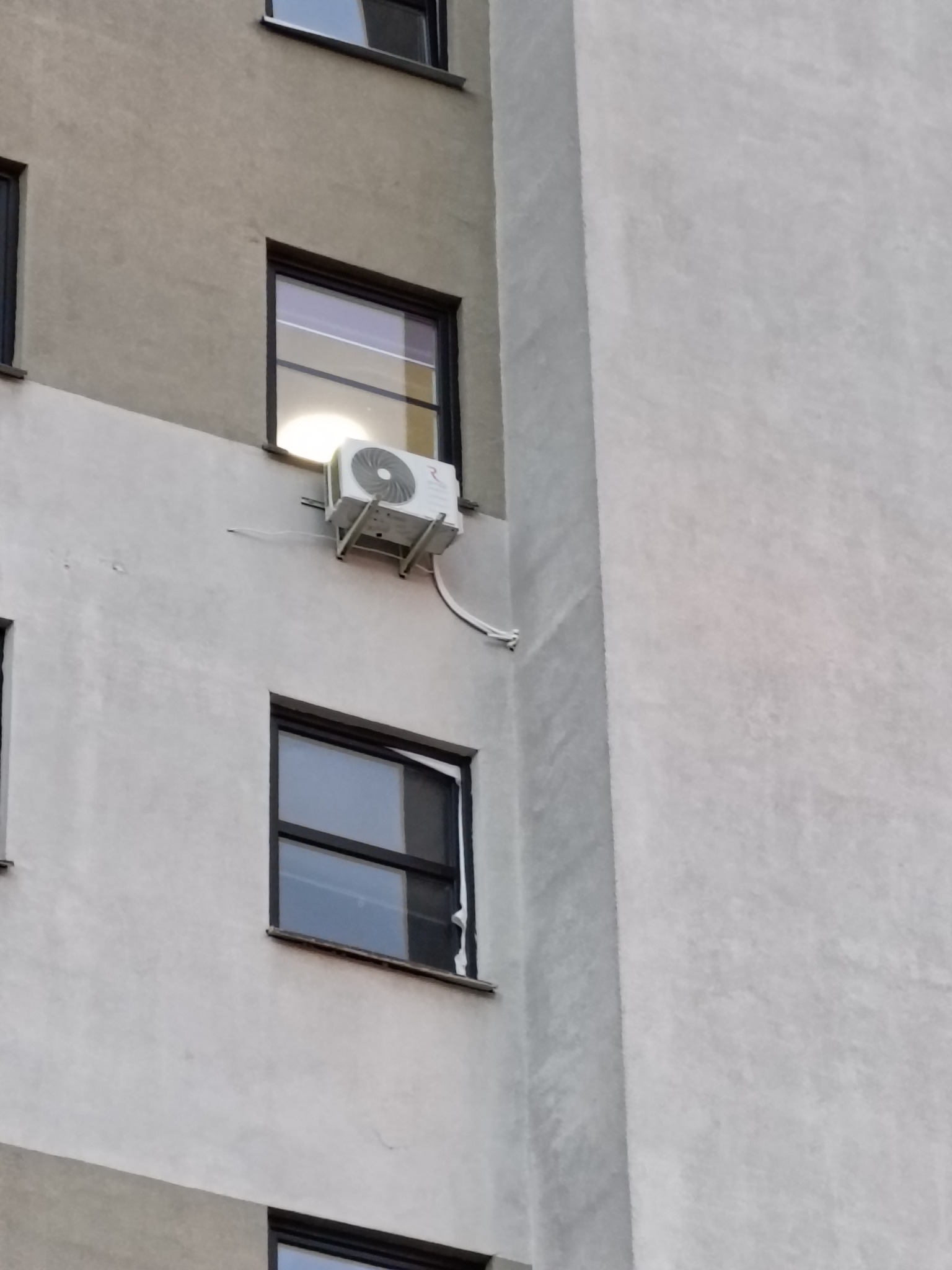 Zewnętrzna jednostka klimatyzatora zamontowana na ścianie budynku mieszkalnego, widoczna z dołu pod kątem, z widocznymi przewodami i fragmentami okien.