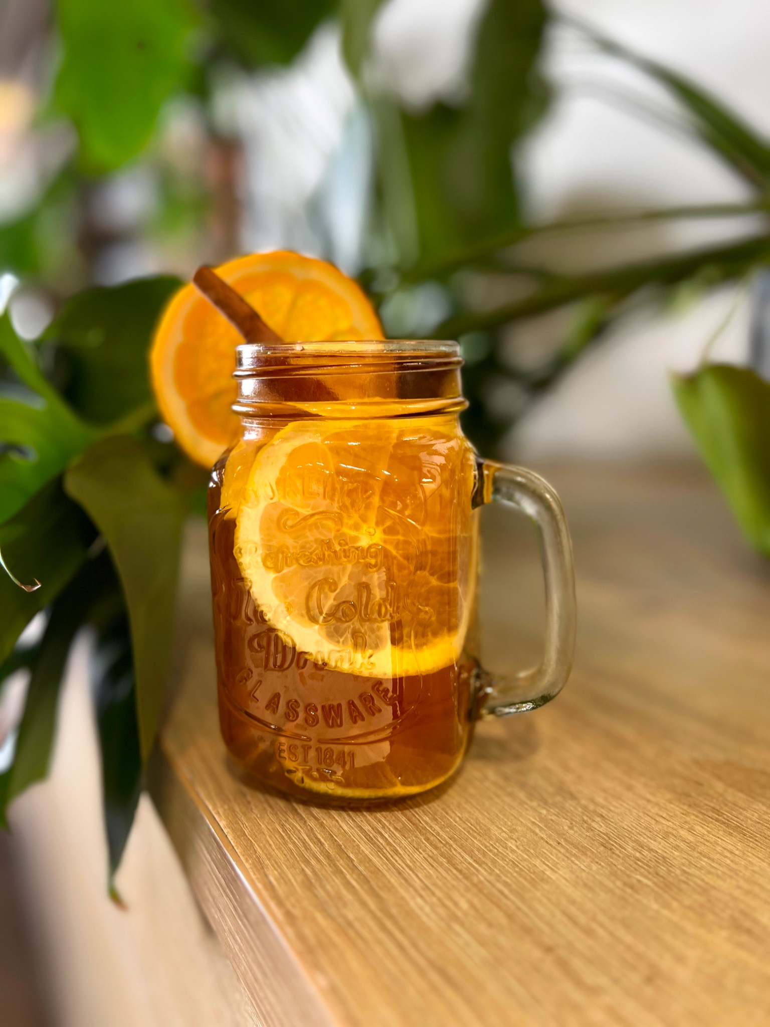 Szklany słoik z uchwytem wypełniony herbatą, plastrami pomarańczy i laską cynamonu, umieszczony na drewnianym blacie na tle zielonych roślin doniczkowych.