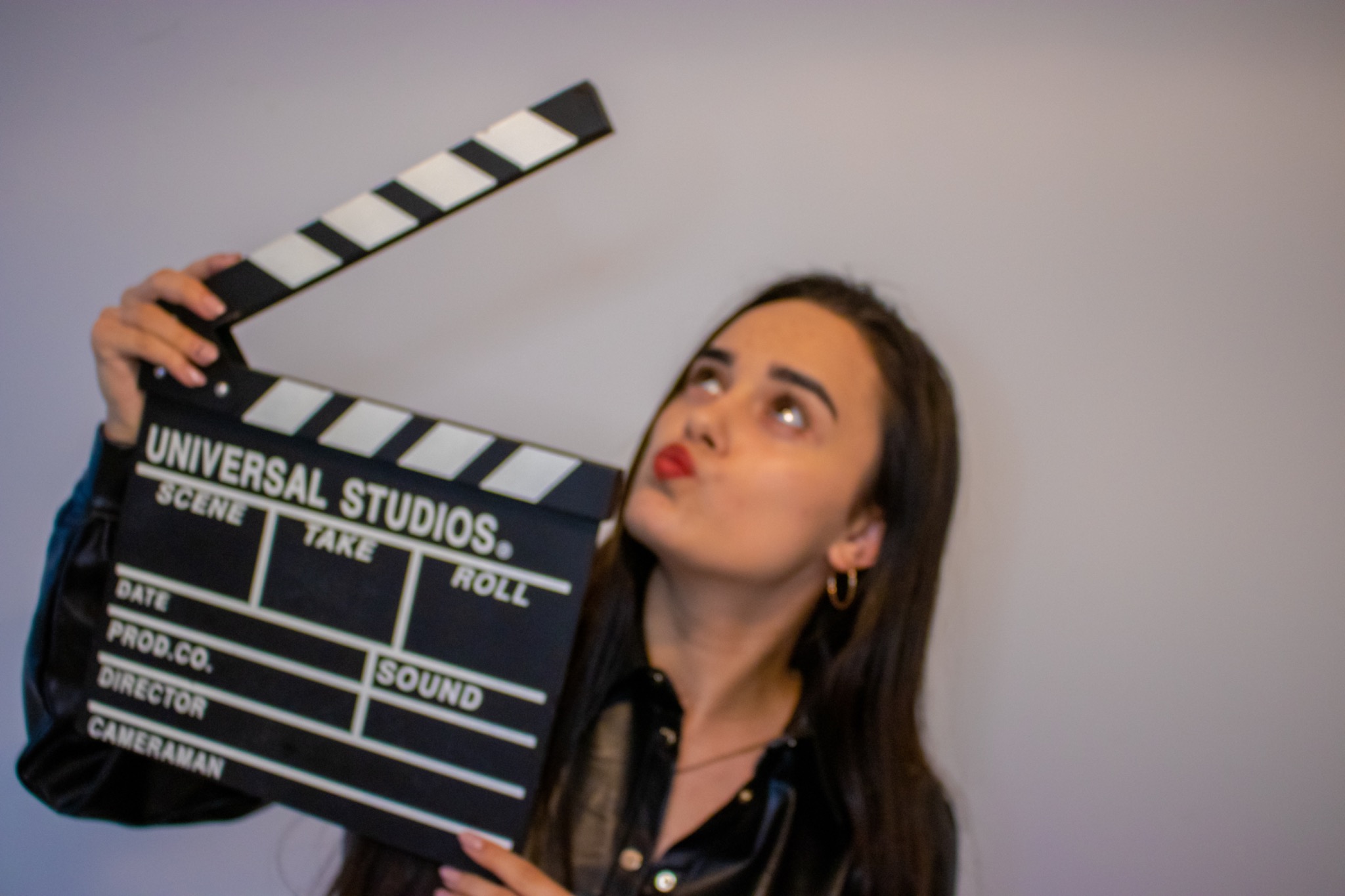 Młoda kobieta w skórzanej kurtce trzyma klaps filmowy Universal Studios, patrząc w górę z zadartymi ustami.