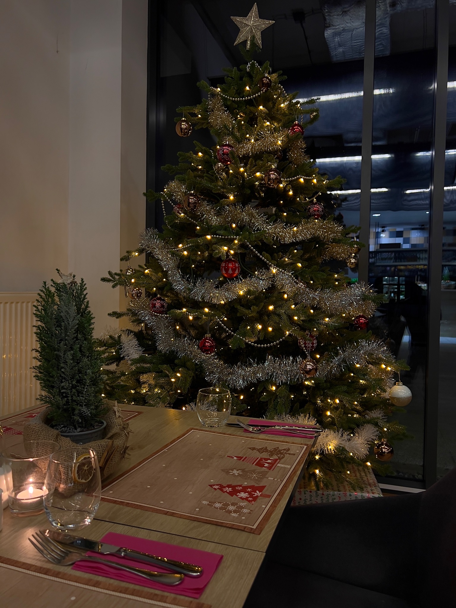 Udekorowany stół z choinką w tle, nakrycie z motywem świątecznym, świece i suszone pomarańcze w szklance, widok z perspektywy siedzącego gościa.