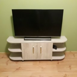 Twoje Meble Na Wymiar - Jasna szafka RTV z półkami i zamkniętą szafką, na której stoi telewizor na tle pistacjowej ściany i drewnianej podłogi.