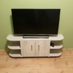 Jasna szafka RTV z półkami i zamkniętą szafką, na której stoi telewizor na tle pistacjowej ściany i drewnianej podłogi.