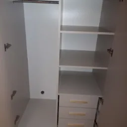 Wnętrze białej szafy z półkami, drążkiem na ubrania i trzema szufladami z poziomymi, złotymi uchwytami, widoczne zawiasy drzwi.