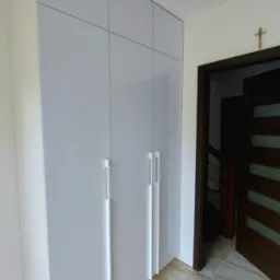 Biała szafa wnękowa z długimi, minimalistycznymi uchwytami, dopasowana do wysokości pomieszczenia, z widoczną podłogą z płytek imitujących marmur.