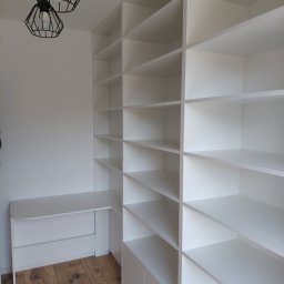 Twoje Meble Na Wymiar - Biała, minimalistyczna szafa wnękowa z biurkiem, oświetlona nowoczesnymi lampami wiszącymi. Jasne wnętrze, drewniana podłoga. Widok z perspektywy użytkownika.