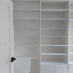 Twoje Meble Na Wymiar - Biała, minimalistyczna szafa wnękowa z biurkiem i półkami, widoczna z perspektywy wnętrza, z otwartymi drzwiami po lewej stronie. Jasne drewno podłogi.