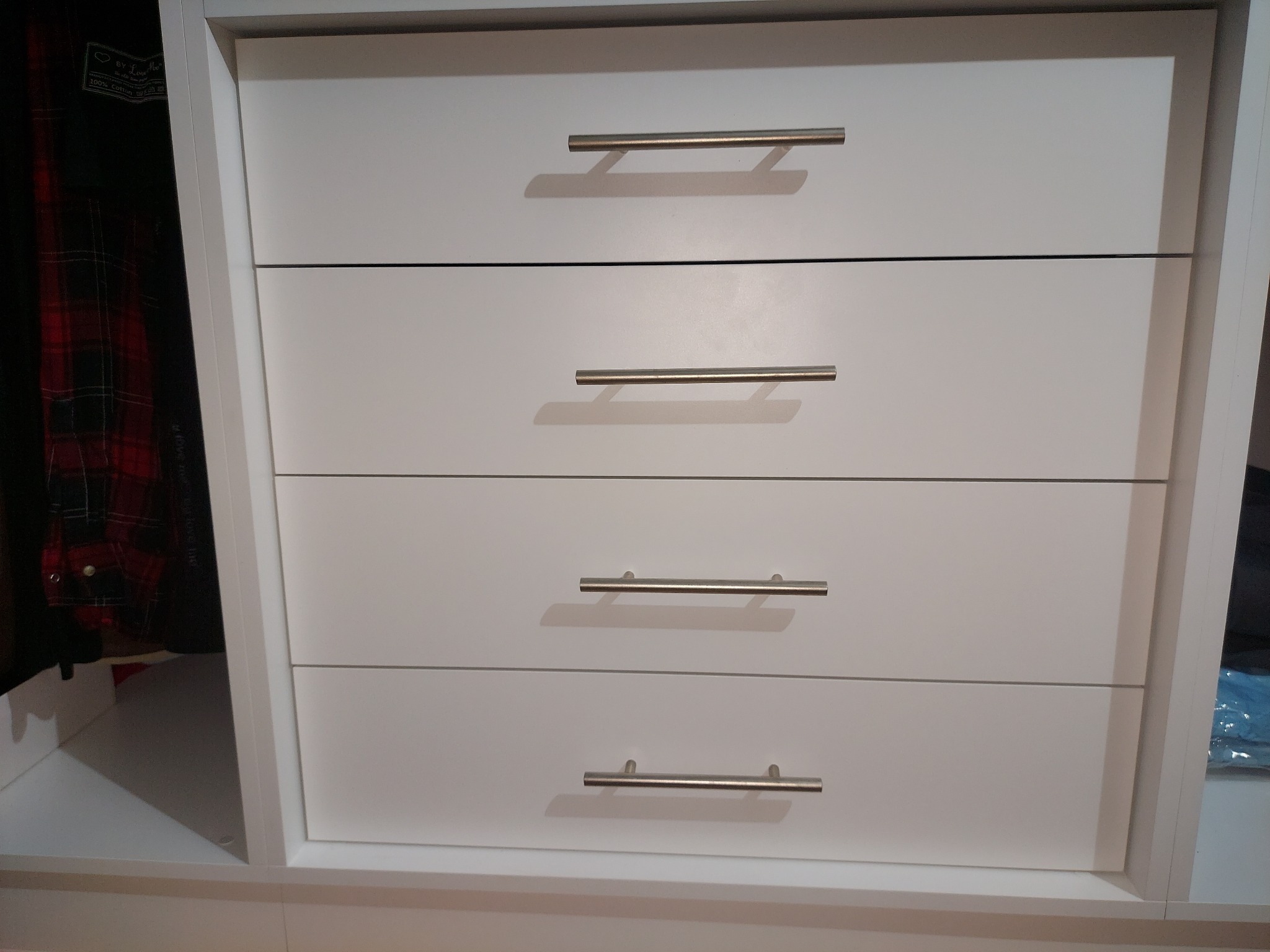 Biała szafa z czterema szufladami i metalowymi uchwytami, widoczna część wnętrza z ubraniami po lewej stronie.