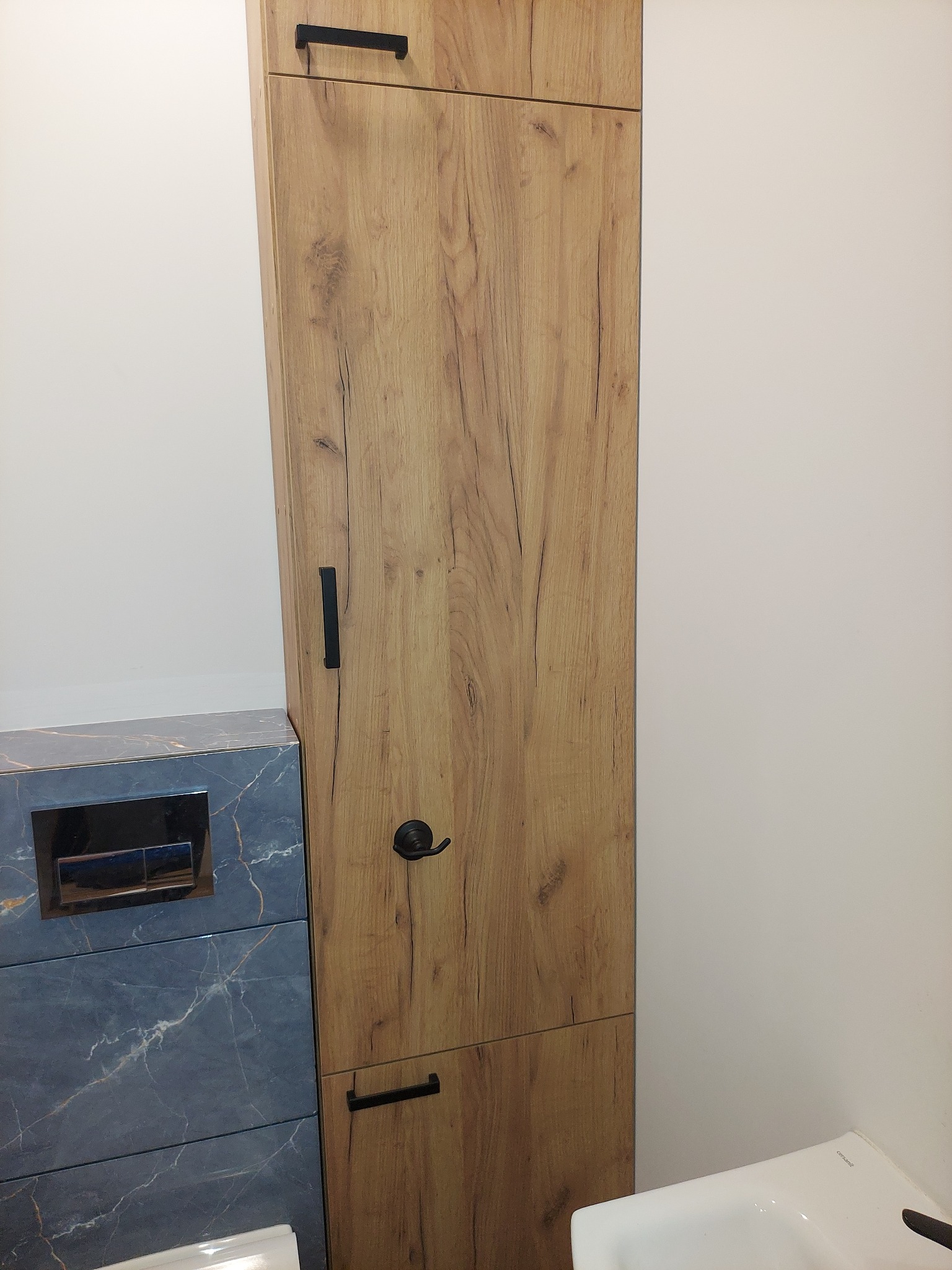 Wysoka, wąska szafka łazienkowa z drewnopodobnym frontem, czarne uchwyty, obok umywalki i zabudowy toalety z marmurkową płytką.