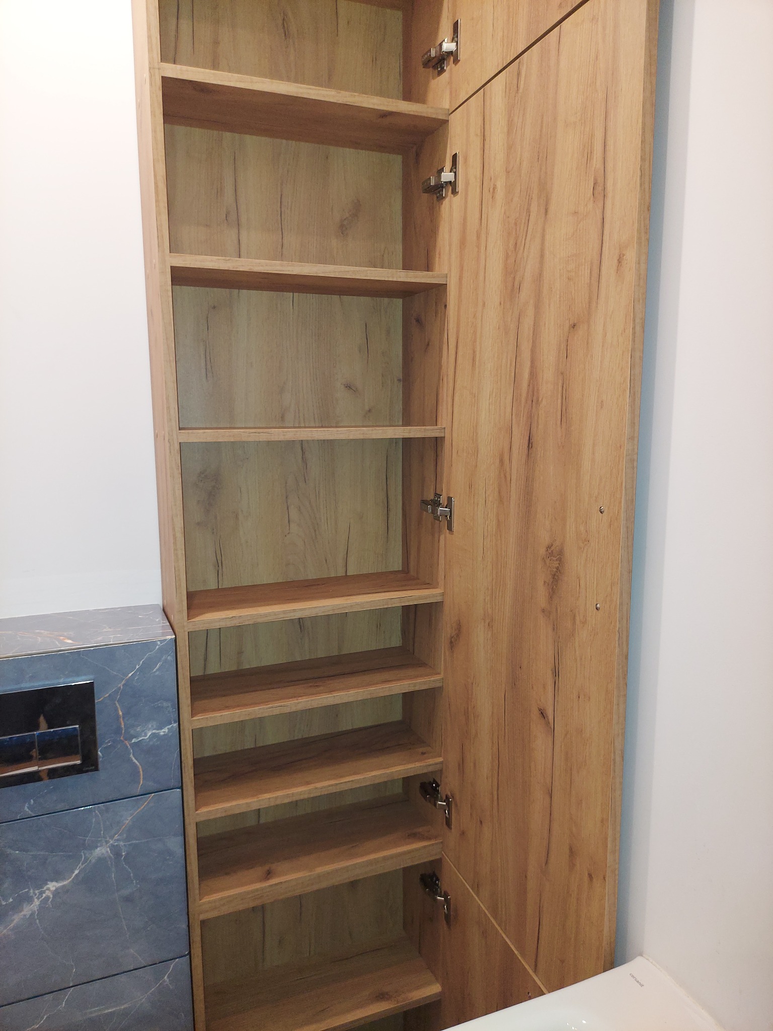 Wysoka, otwarta szafa z drewnianymi półkami, zamontowana w łazience obok zabudowy WC z marmurowym wzorem.