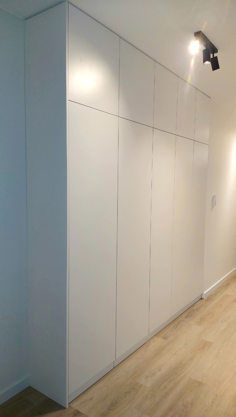 Biała szafa wnękowa z gładkimi frontami, oświetlenie sufitowe punktowe, jasna podłoga panelowa.