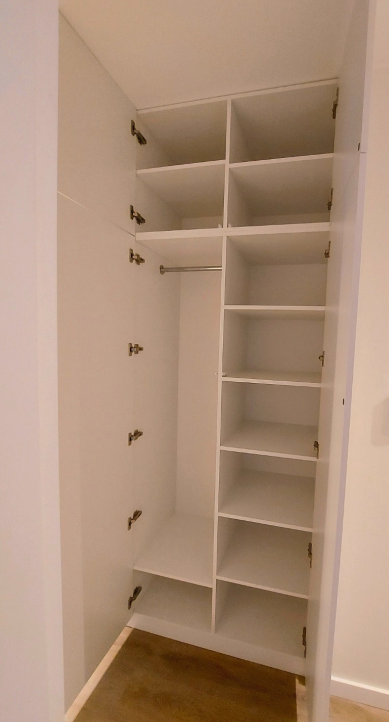 Wnętrze białej szafy z półkami i drążkiem na ubrania, widok z otwartymi drzwiami, widoczne zawiasy.
