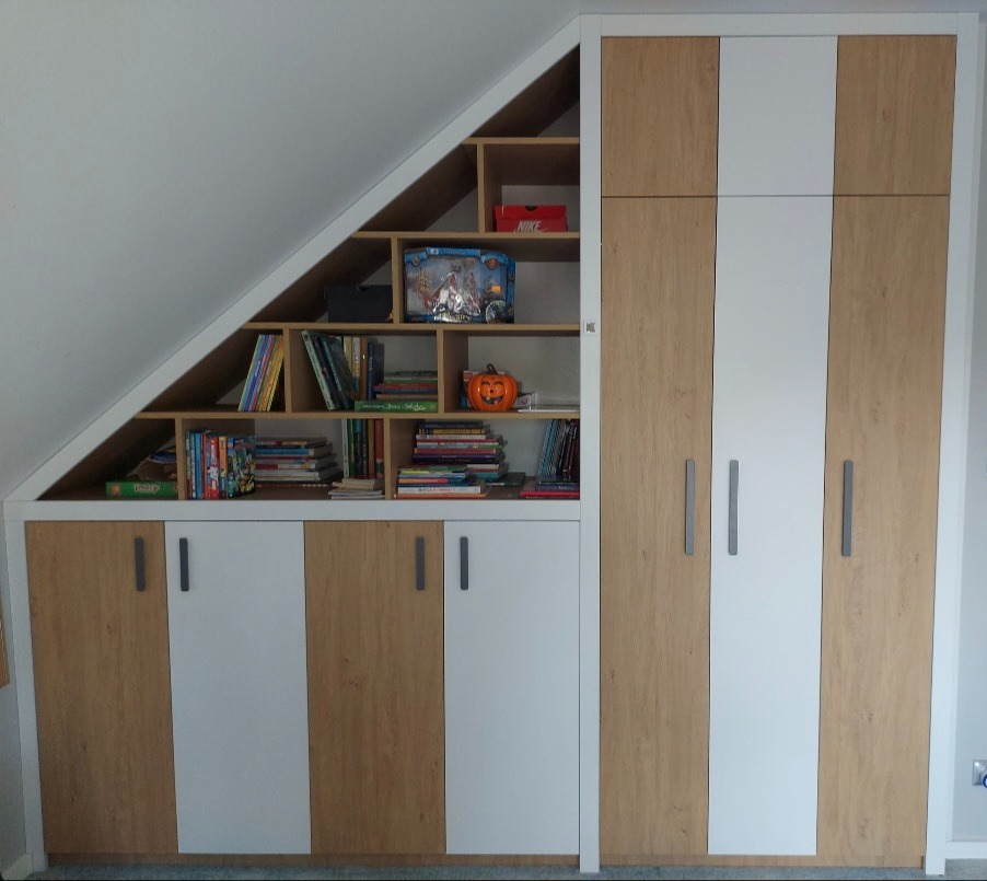 Szafa wnękowa z półkami pod skosem dachu, fronty biało-drewniane, na półkach książki, zabawki i dekoracja dyni na Halloween.