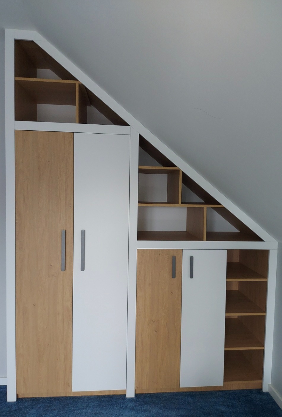 Szafa wnękowa na poddaszu z białymi i drewnopodobnymi frontami, z otwartymi półkami w trójkątnej przestrzeni pod skosem dachu.