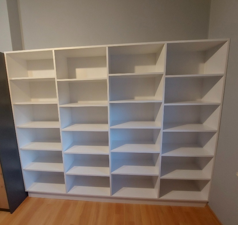 Biała, duża szafa wnękowa z wieloma półkami, bez zawartości, na tle jasnej ściany i drewnianej podłogi.