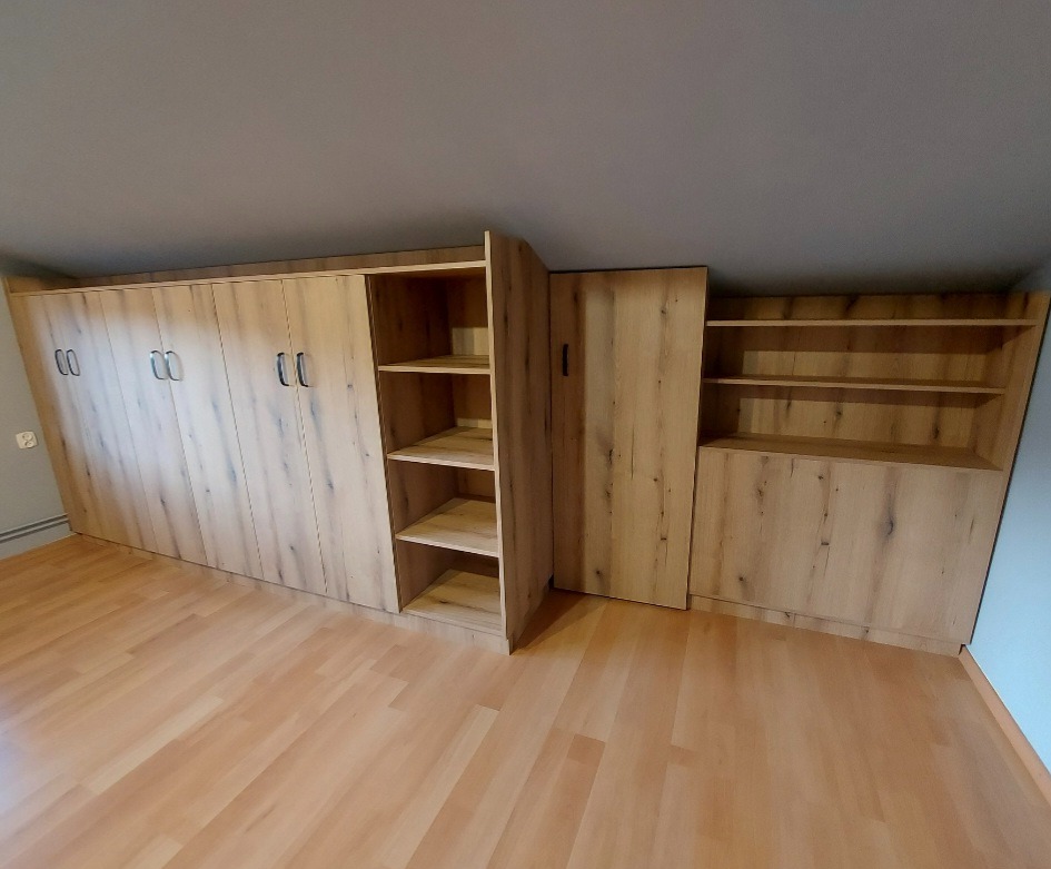 Zabudowa pod skosem z jasnego drewna: szafa z frontami na zawiasach, regał z półkami i szafka z półkami w górnej części, na podłodze panele