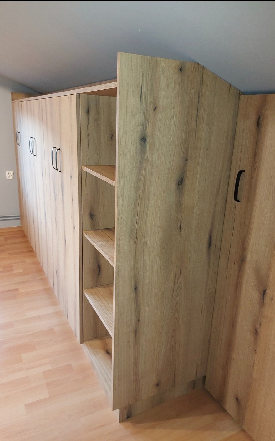 Długa szafa wnękowa z otwartymi półkami z płyty imitującej jasne drewno, z czarnymi uchwytami, dopasowana do skosu sufitu.