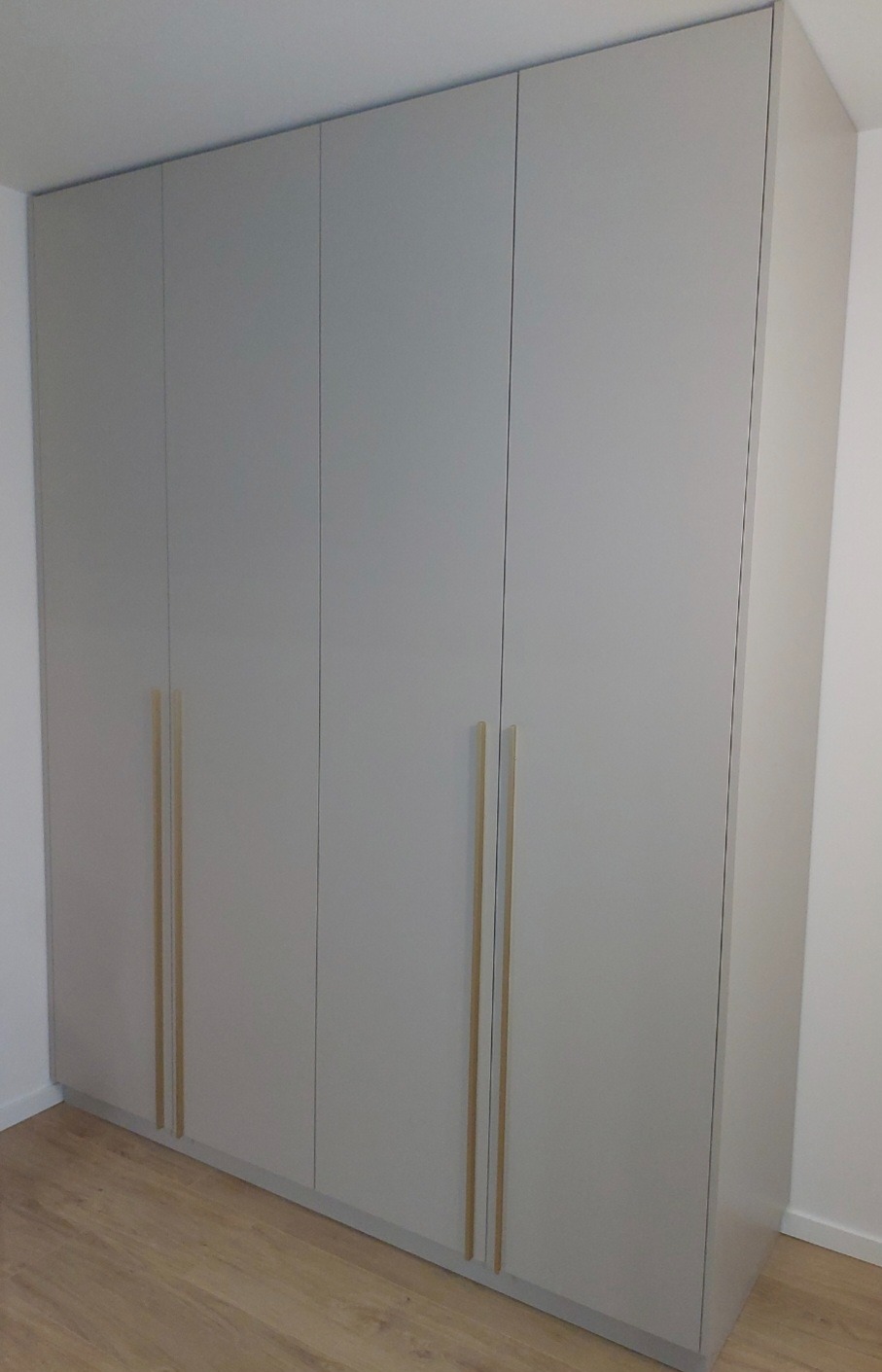 Czterodrzwiowa, szara szafa wnękowa z długimi, pionowymi, jasnymi uchwytami na drewnianej podłodze.