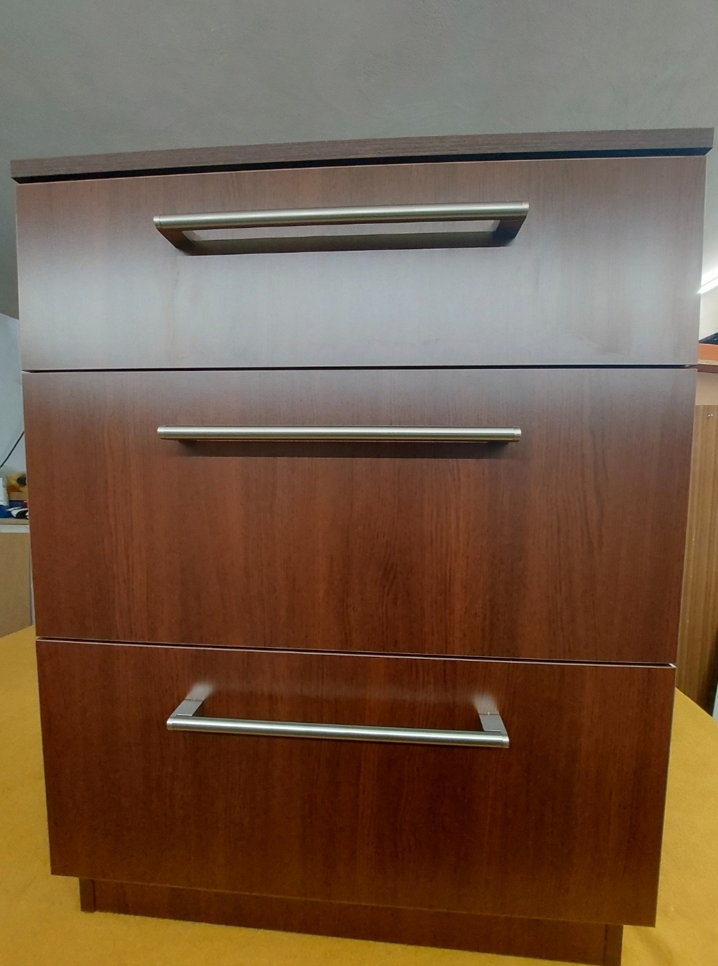 Komoda z trzema szufladami o wykończeniu imitującym ciemne drewno, z prostymi, metalowymi uchwytami.