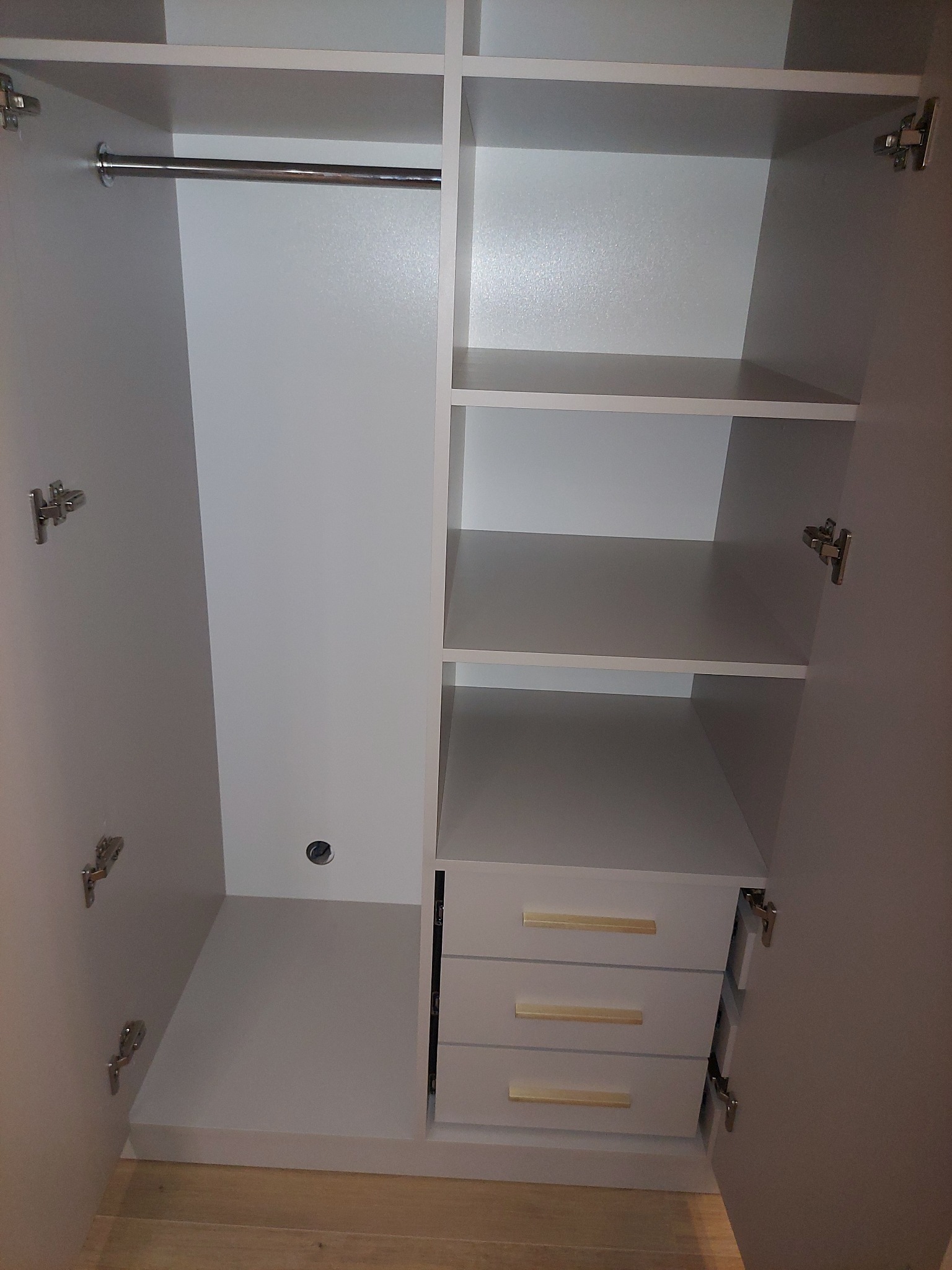 Wnętrze białej szafy z półkami, drążkiem na ubrania i trzema szufladami z poziomymi, złotymi uchwytami, widoczne zawiasy drzwi.