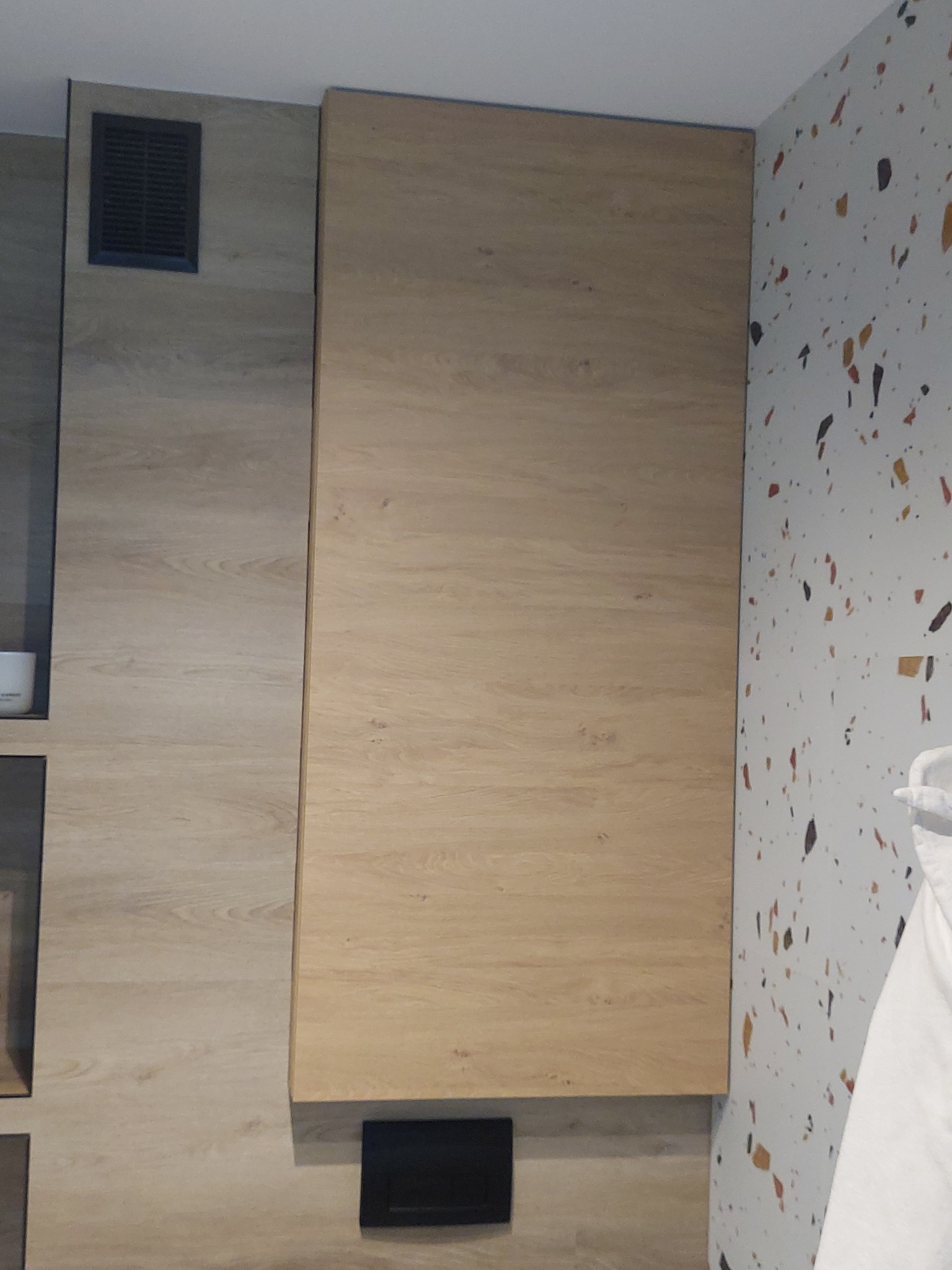 Zamknięta szafa wnękowa z drewnianymi frontami, z widoczną kratką wentylacyjną w górnej części i czarnym elementem w dolnej części, na tle ściany z wzorem lastryko.