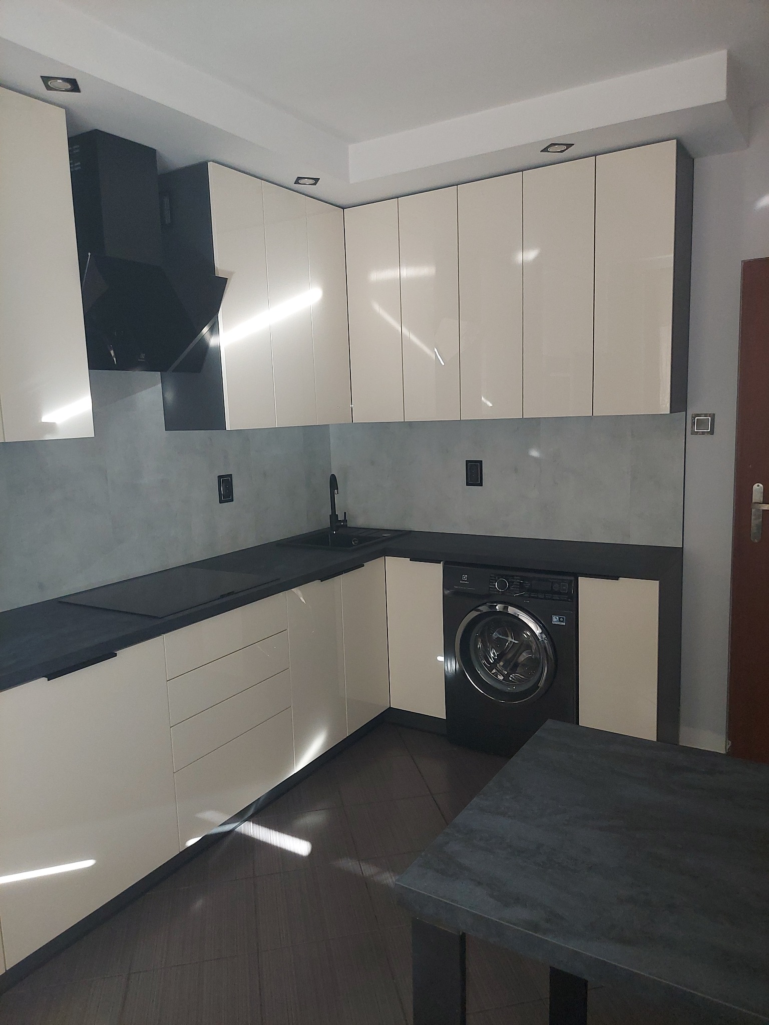 Jasna kuchnia z zabudową pod sufit i czarnym blatem, z pralką w zabudowie i szarą ścianą nad blatem.