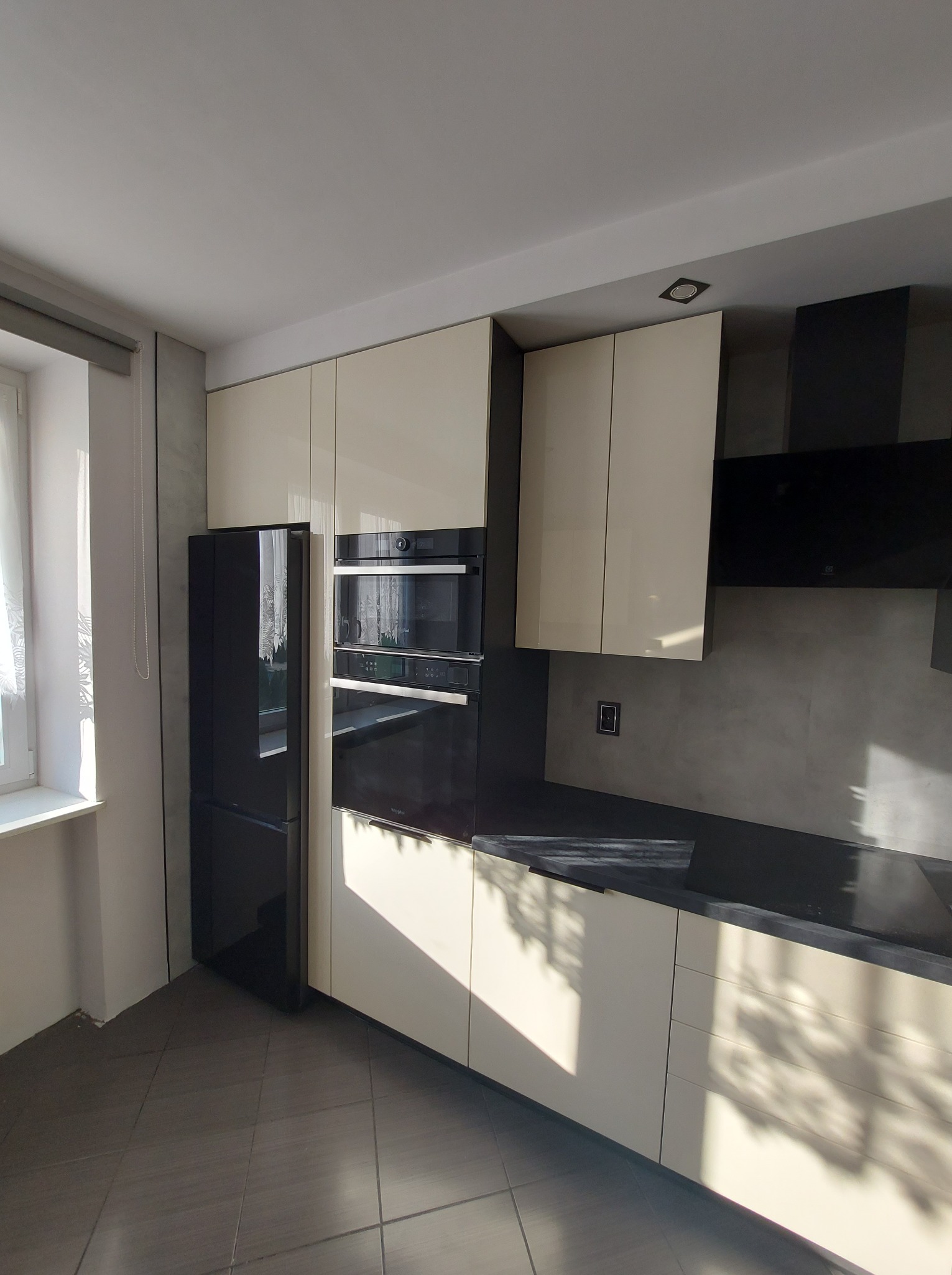 Nowoczesna kuchnia z beżowymi szafkami, czarnym blatem i sprzętem AGD w zabudowie, oświetlona naturalnym światłem padającym z okna.