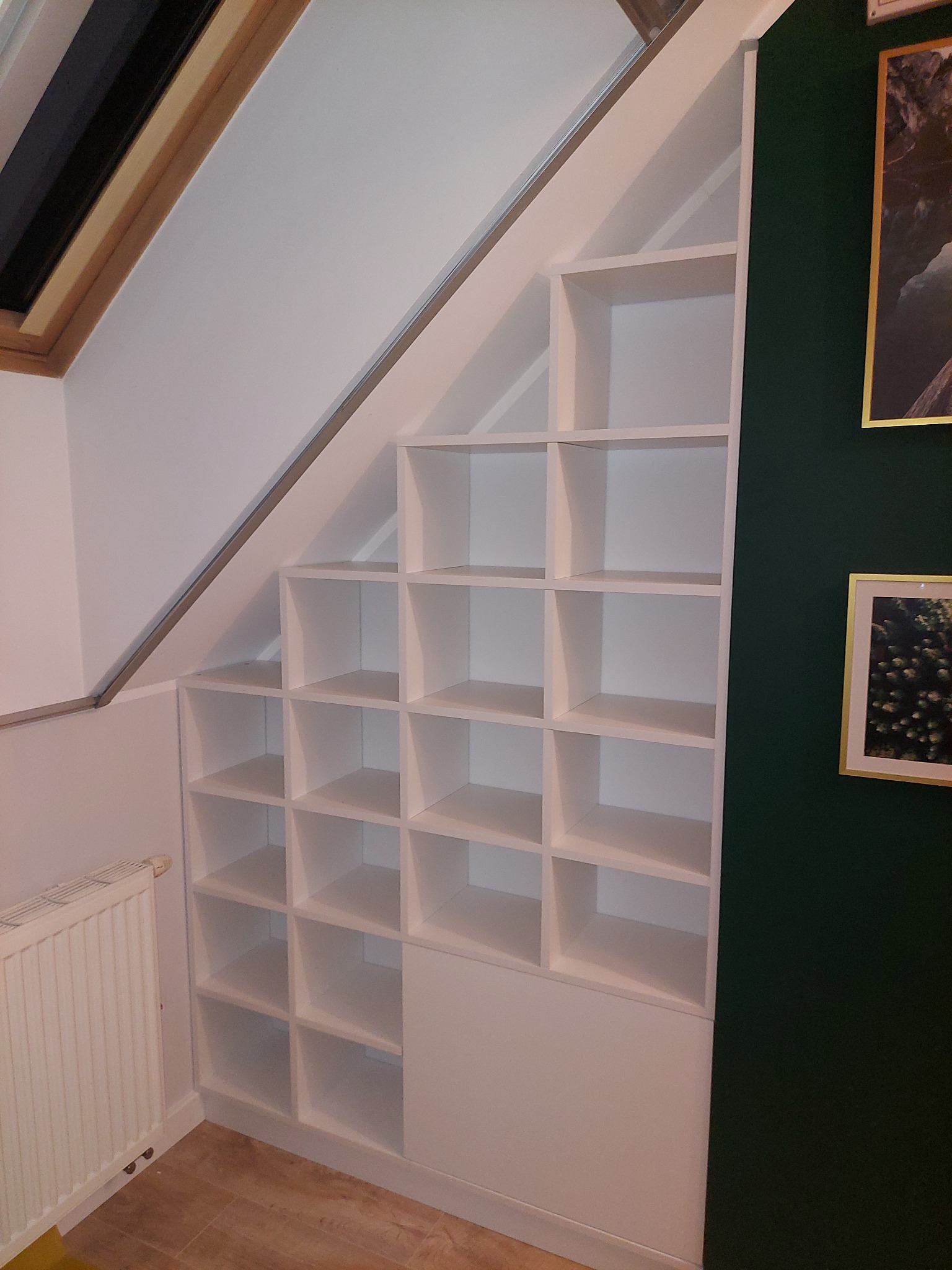 Biała, modułowa szafa wnękowa z otwartymi półkami, dopasowana do skosu poddasza, z grzejnikiem obok i zieloną ścianą w tle.